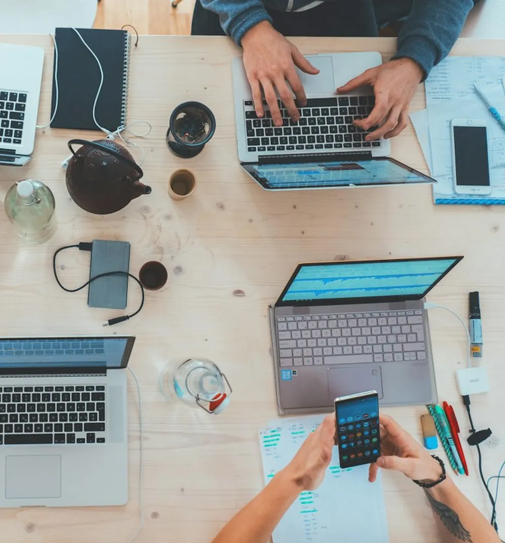 Manos revisando un teléfono y gráficos junto a portátiles en una mesa de trabajo